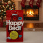 Red 'Happy Bear' package in front of a festive fireplace and Christmas tree.